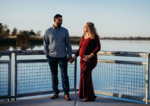 Pregnant woman in a red dress holding hands with her partner on a lakeside boardwalk