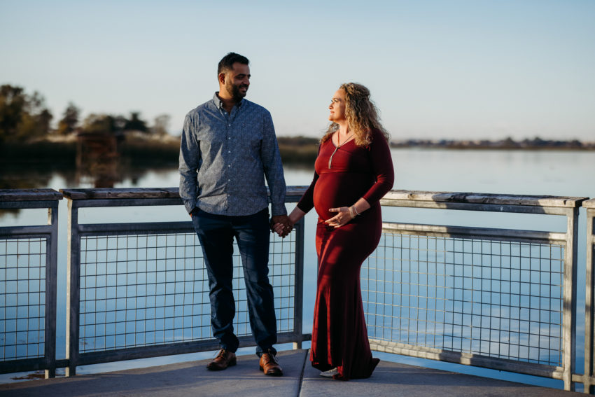 Pregnant woman in a red dress holding hands with her partner on a lakeside boardwalk